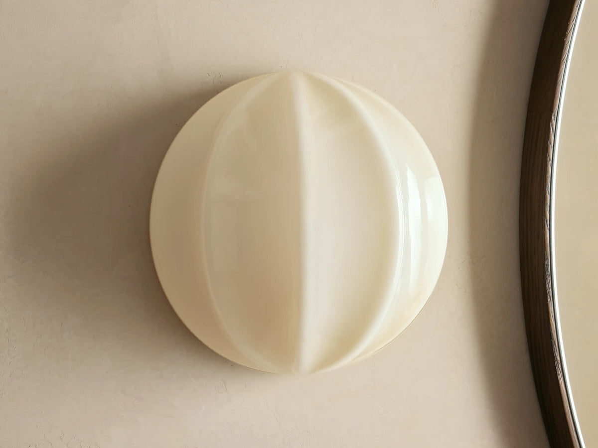 Agnes Clamshell Sconce: Spherical cream paneled glass sconce, glowing warmly, mounted on a light wall next to a dark mirror.