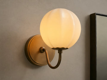 Agnes Gooseneck Sconce with antique brass base, curved arm, and glowing paneled cream glass globe shade mounted on a wall.