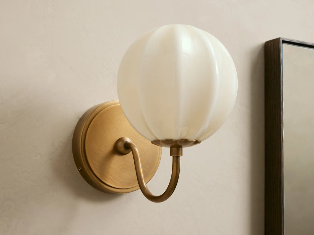 Agnes Gooseneck Sconce with antique brass base, curved arm, and glowing paneled cream glass globe shade mounted on a wall.