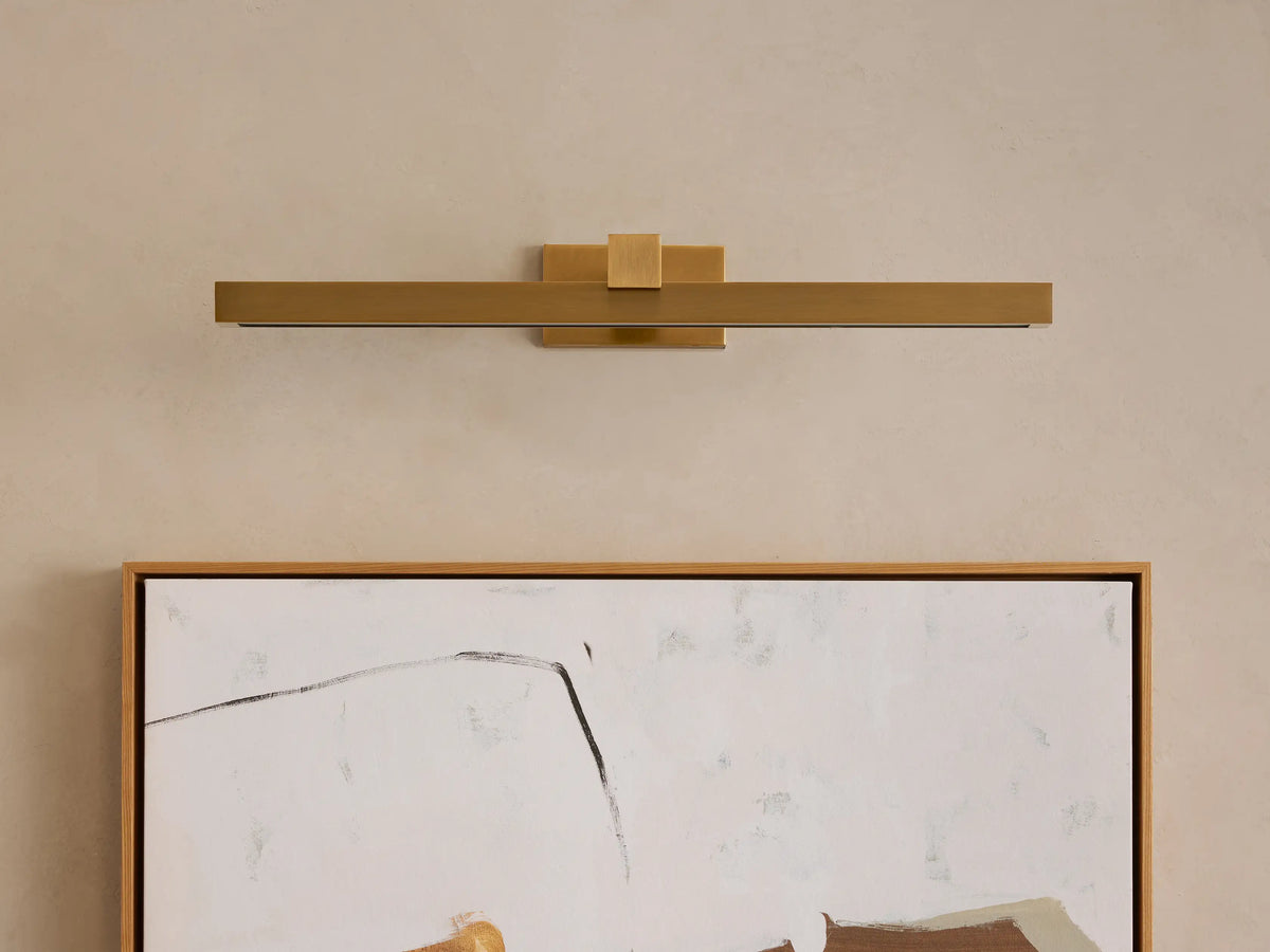 Corinth Picture Light Sconce, brushed brass, rectangular bar design with illuminated LED strip, mounted horizontally above abstract art.