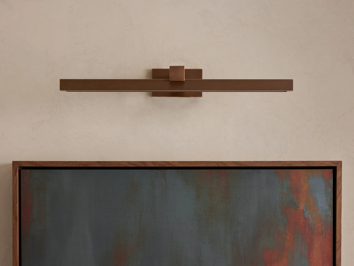 Corinth Picture Light Sconce, a long, horizontal bronze fixture on a light wall, casting warm LED light onto a framed abstract painting.