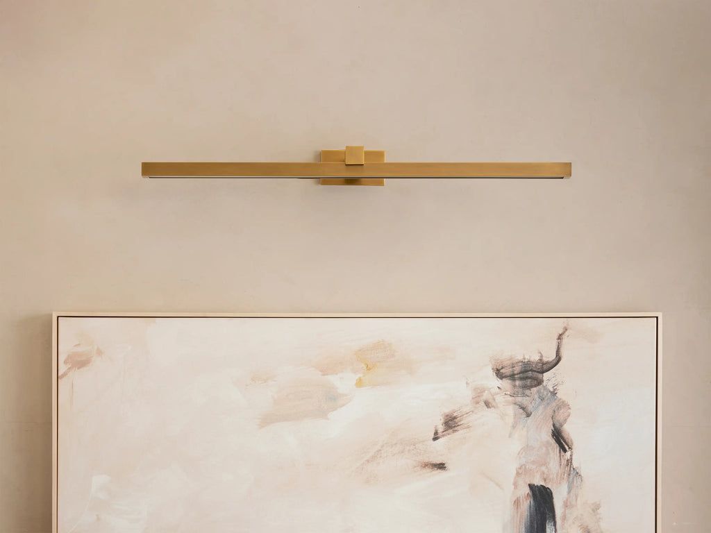 Corinth Picture Light Sconce in brass finish, mounted horizontally on a beige wall, illuminating an abstract painting below.