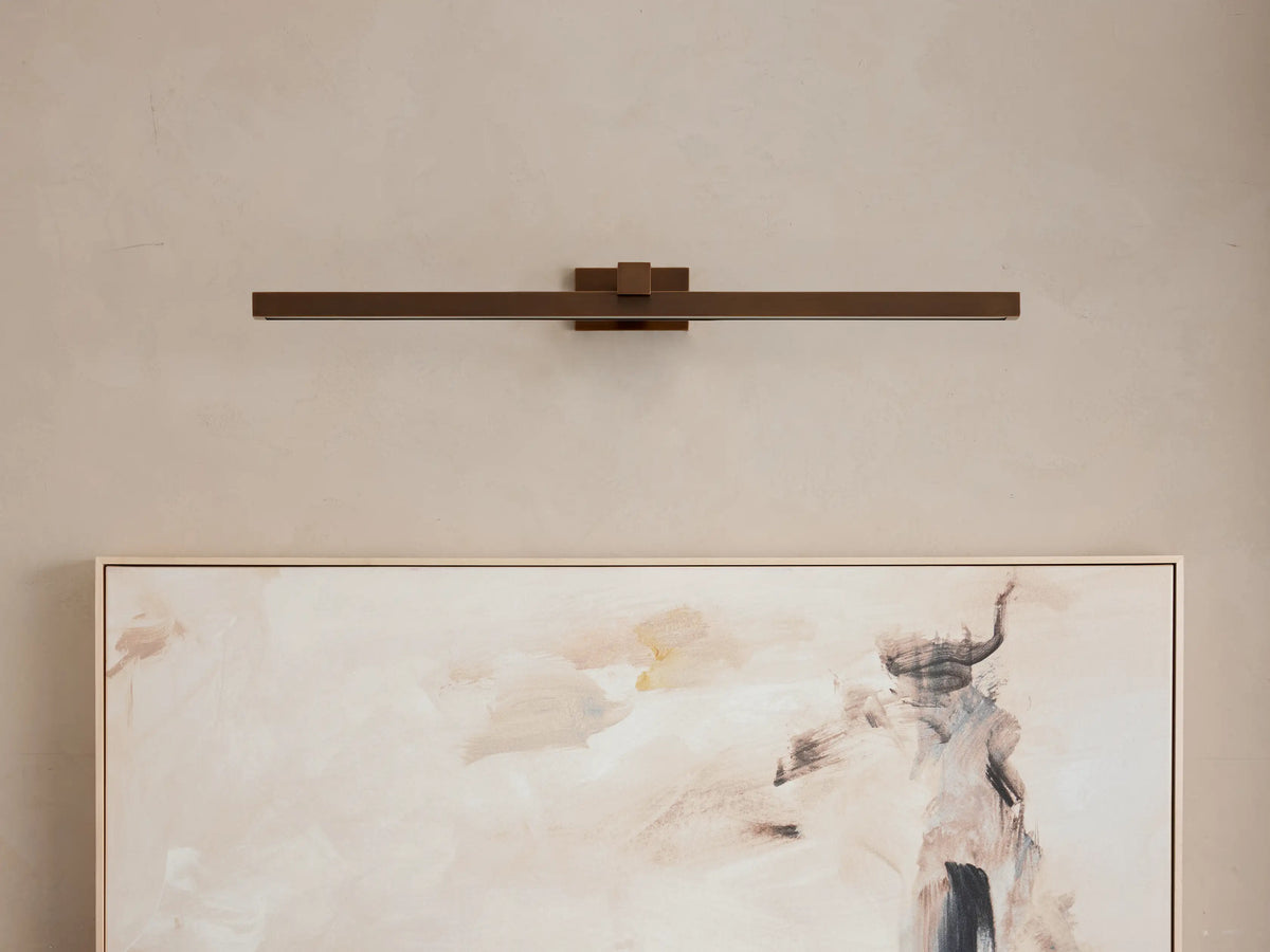 Dark bronze Corinth Picture Light Sconce, horizontal, emitting warm LED light downward onto an abstract painting on a beige wall.