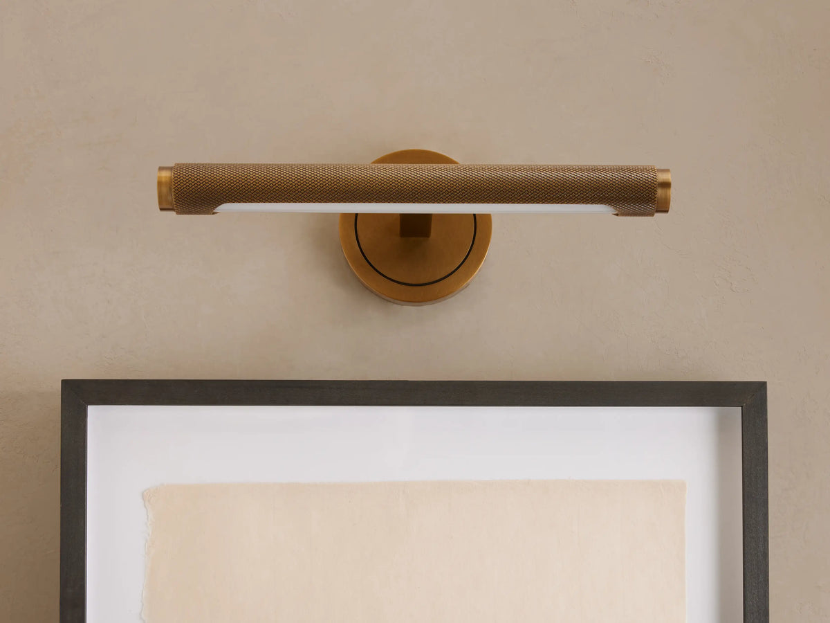 Antique brass Monroe Picture Light Sconce with knurled texture, mounted on a beige wall, illuminating framed art.