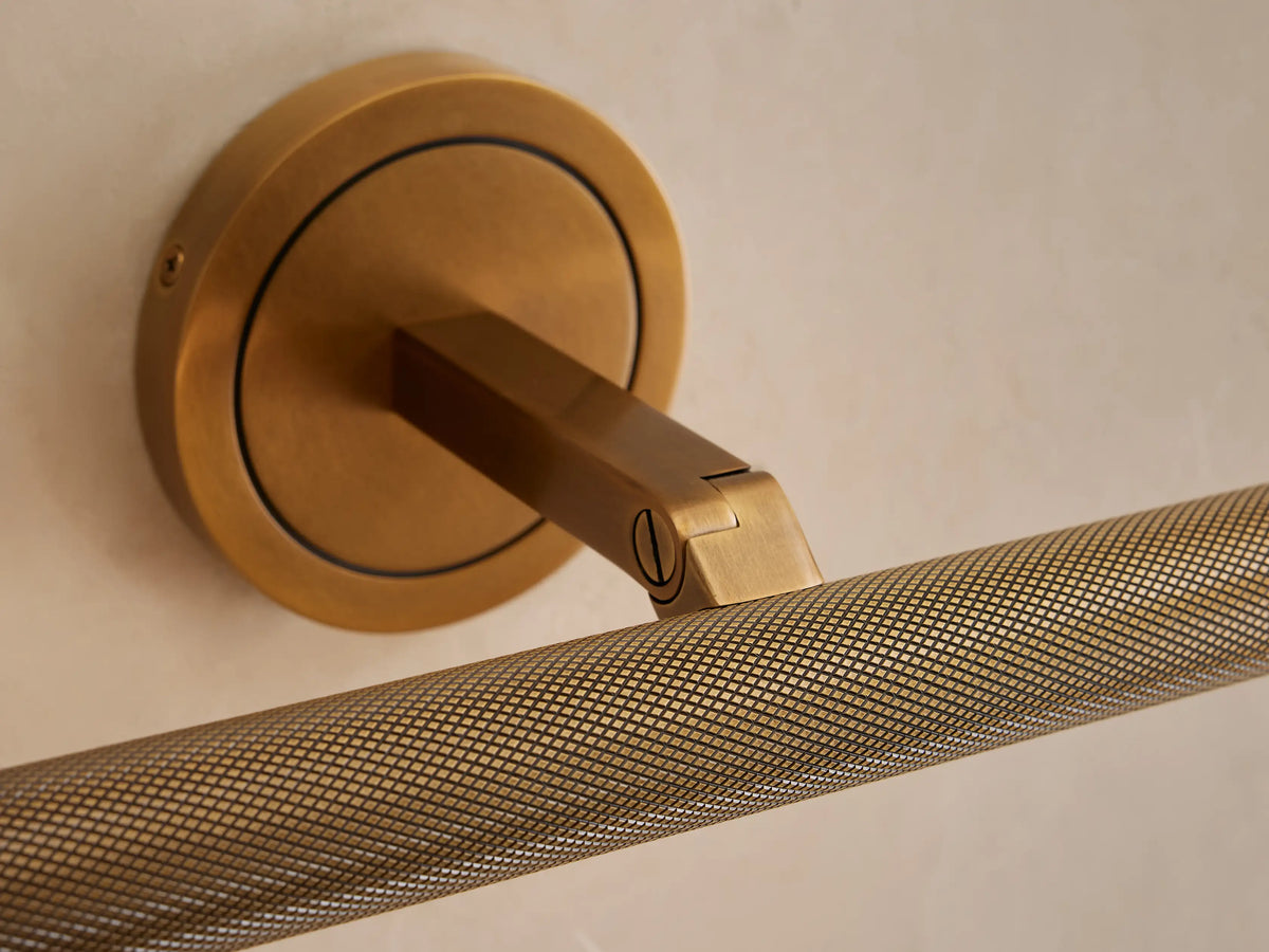 Close-up of an antique brass Monroe Picture Light Sconce mounted on a light wall, featuring a knurled textured light bar.