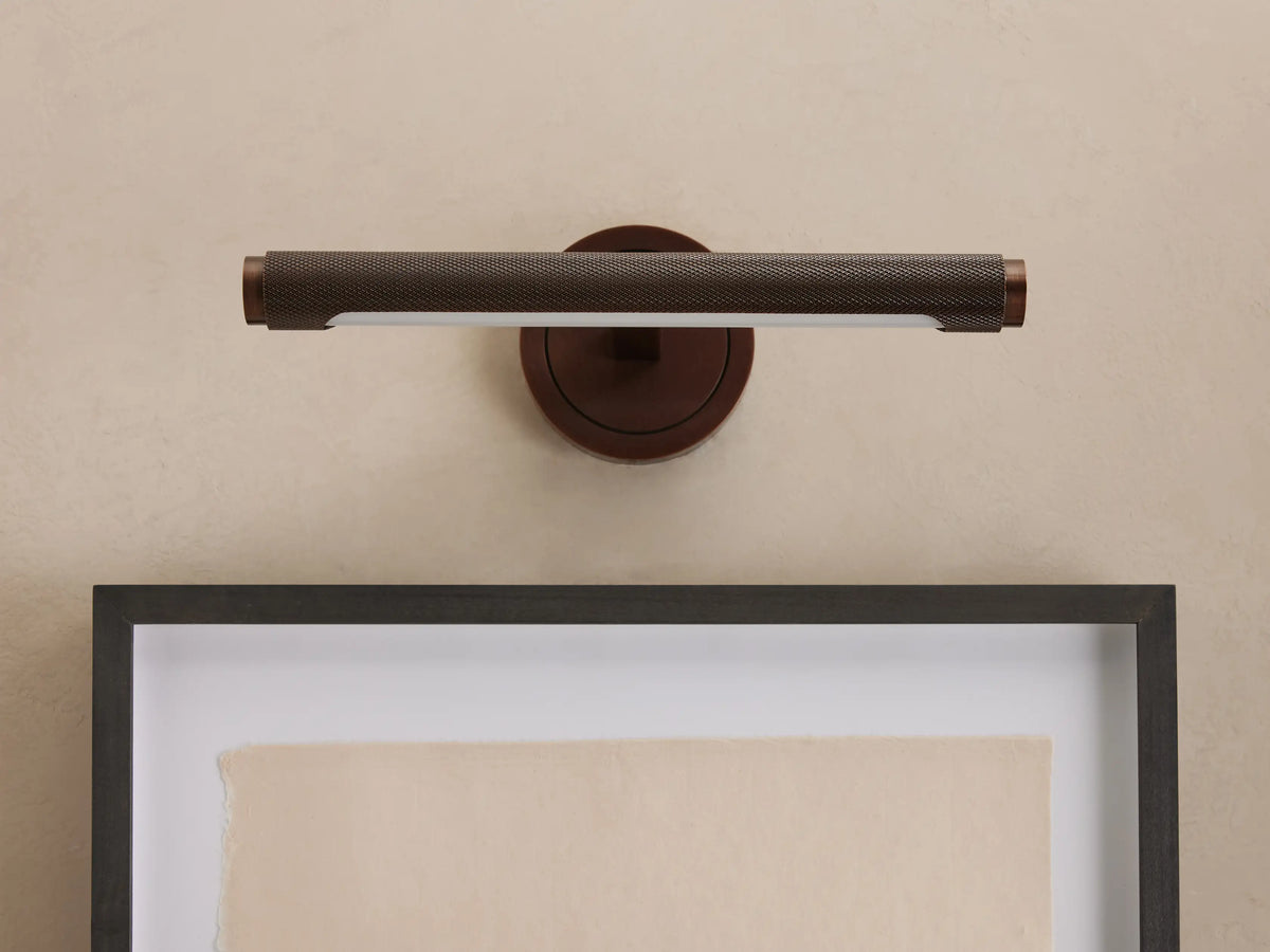 Monroe Picture Light Sconce in antique brass with knurled texture, mounted on a light wall above a dark-framed artwork.