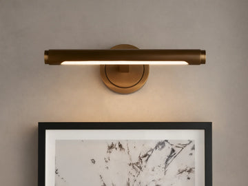 Monroe Picture Light Sconce in antique brass, emitting warm LED light over a framed abstract artwork on a light-colored wall.