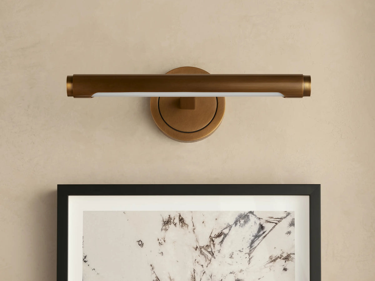 Antique brass Monroe Picture Light Sconce on a beige wall, its integrated LED illuminating a framed abstract artwork below.