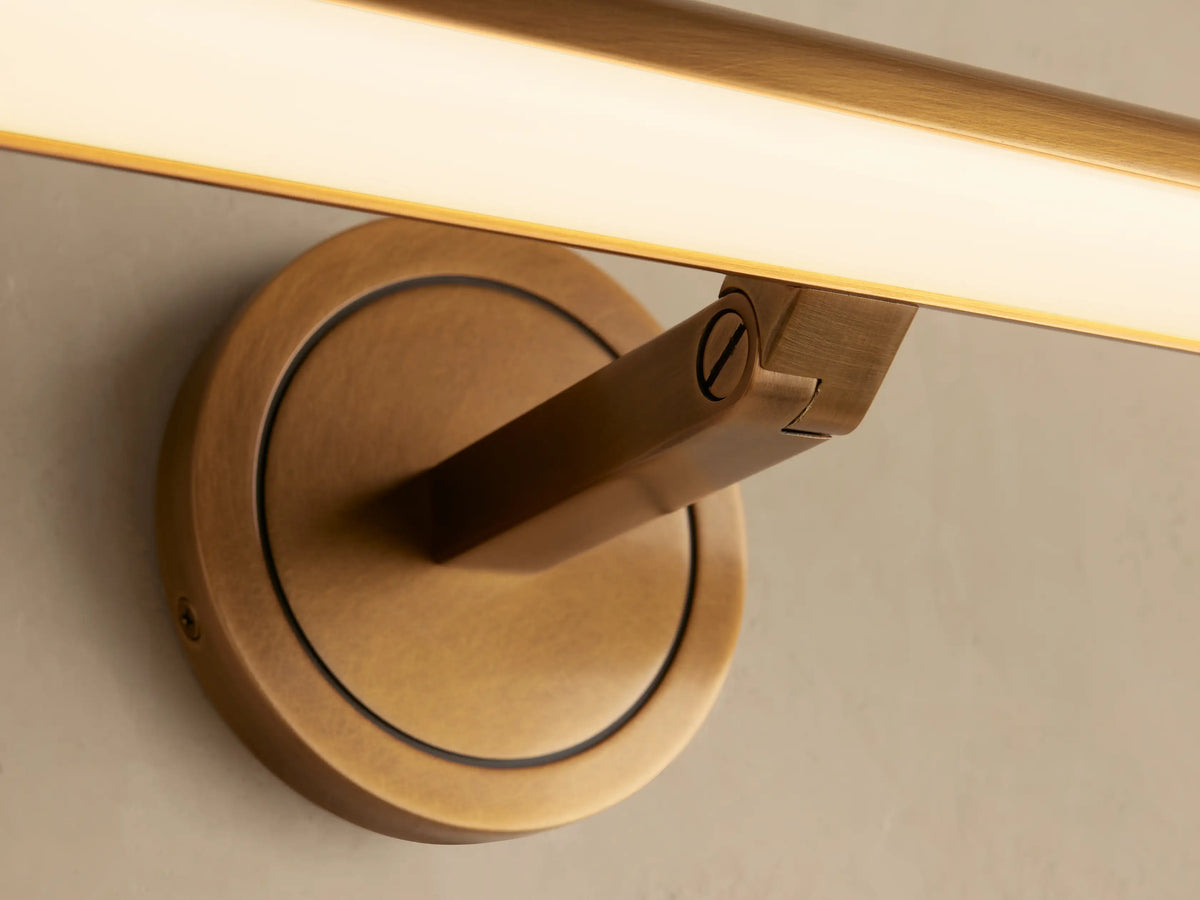 Close-up of antique brass Monroe Picture Light Sconce. Round wall mount, angled arm, and illuminated LED bar with warm light.