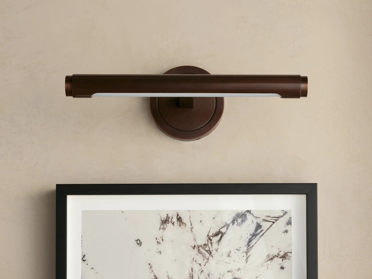 Monroe Picture Light Sconce, antique brass, linear, with a white acrylic diffuser, mounted on a wall above framed art.