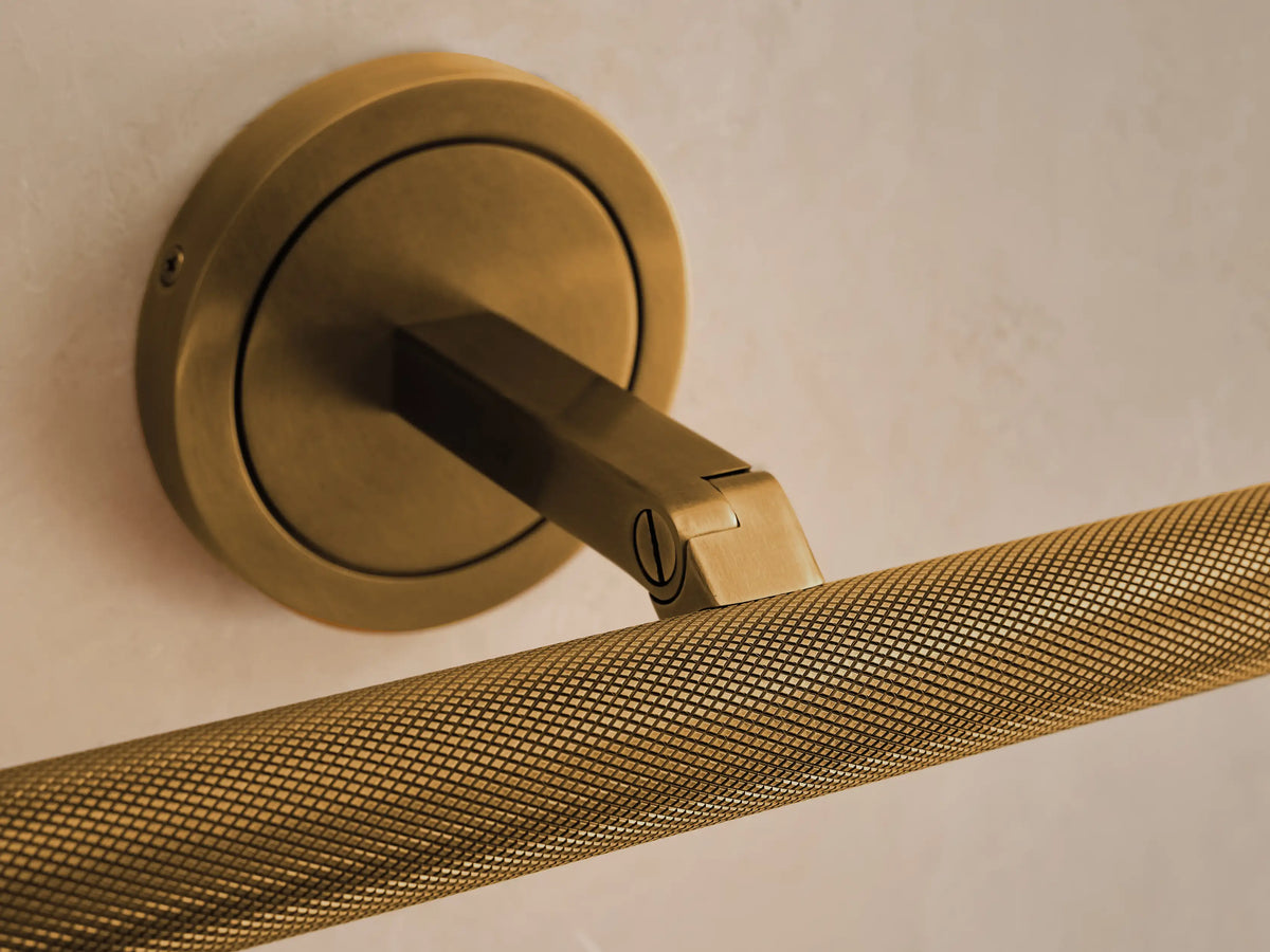 Antique brass Monroe Picture Light Sconce with round base, arm, and horizontal knurled light bar, against a cream wall.