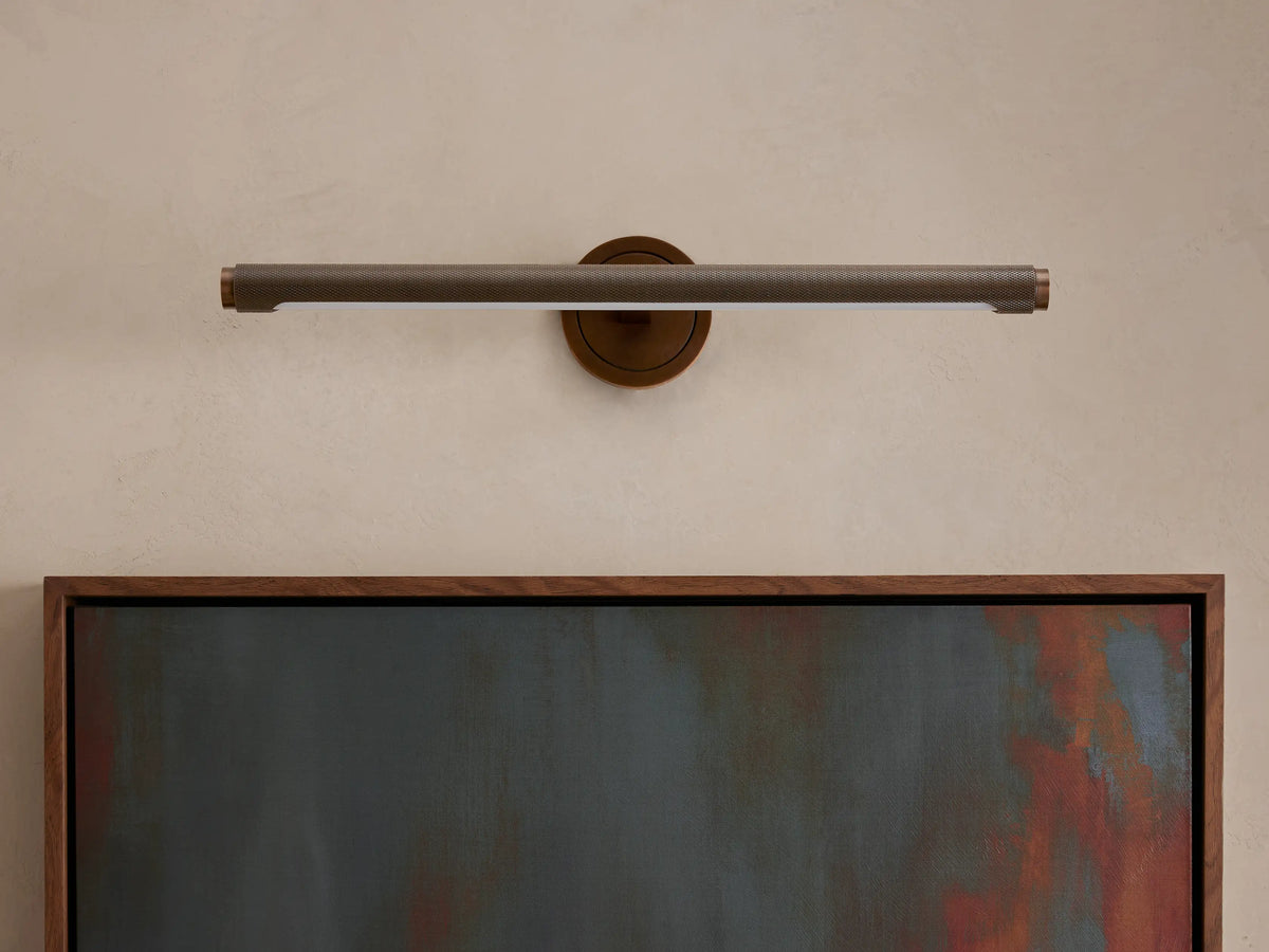 Monroe Picture Light Sconce, horizontal, knurled antique brass, lighted, above a framed abstract painting.