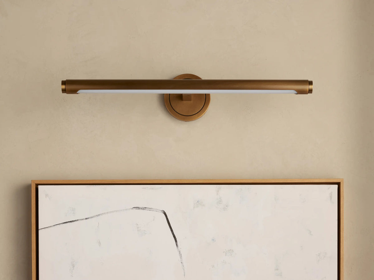 Monroe Picture Light Sconce in antique brass finish, horizontal bar with LED strip, mounted on a beige wall above abstract art.