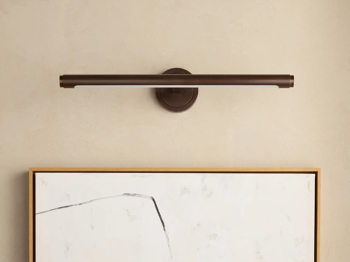 Monroe Picture Light Sconce in antique brass, mounted on a beige wall, illuminating an abstract artwork with a wood frame.