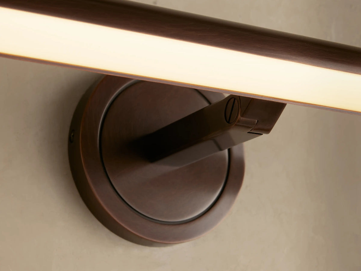 Close-up of Monroe Picture Light Sconce in antique brass. The arm extends from a circular base, emitting warm LED light on a beige wall.