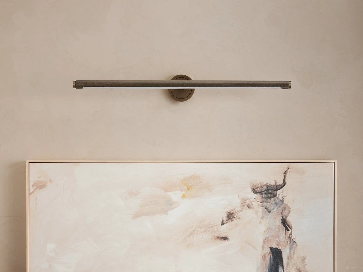Monroe Picture Light Sconce in antique brass, a horizontal bar-style light mounted on a beige wall above an abstract painting.