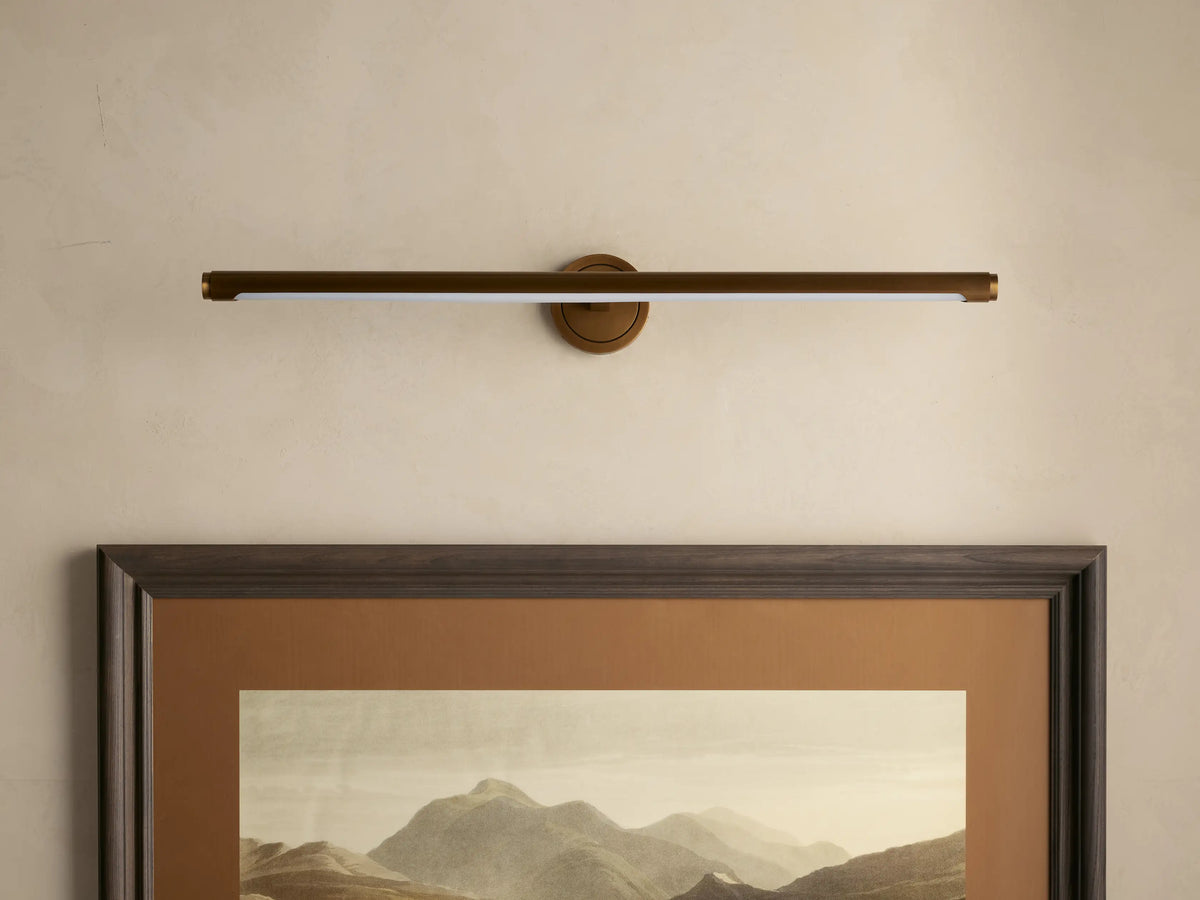 Antique brass Monroe Picture Light Sconce with a soft LED glow over a framed mountain landscape on a beige wall.