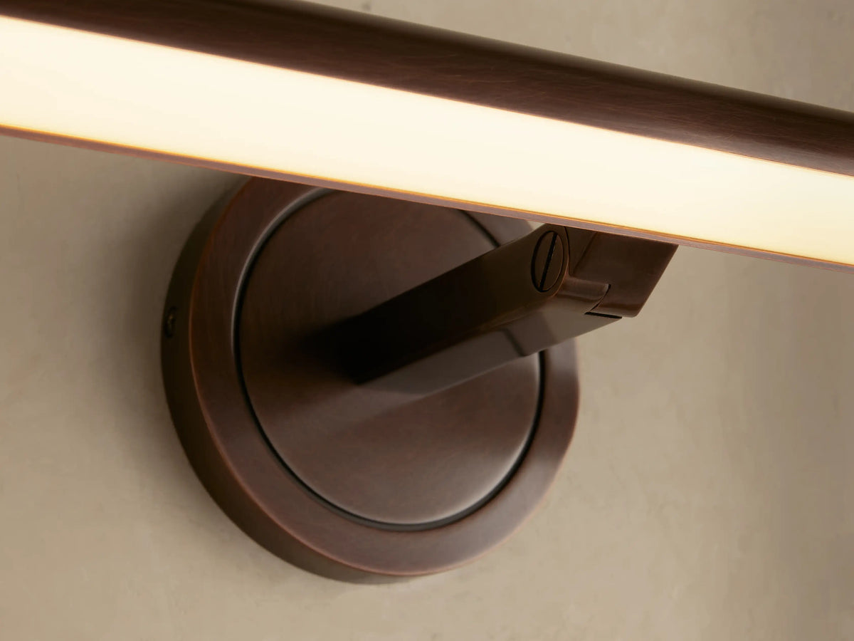 Antique brass Monroe Picture Light Sconce, mounted on a light wall, with its acrylic diffuser glowing with warm LED light.