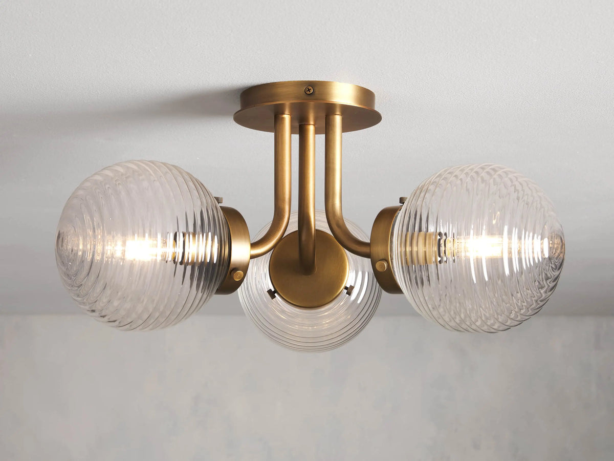 Sayre Flush Mount with burnished brass finish, features three ribbed glass globes attached to a central steel fixture against a light background.