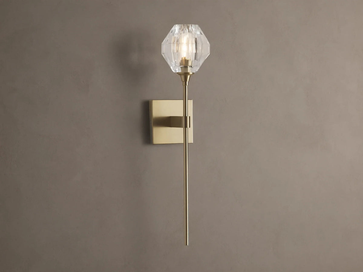 Amoroso Sconce: brass-plated wall light with long rod and illuminated faceted clear crystal shade on a brown wall.