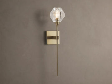 Amoroso Sconce: brass-plated wall light with long rod and illuminated faceted clear crystal shade on a brown wall.