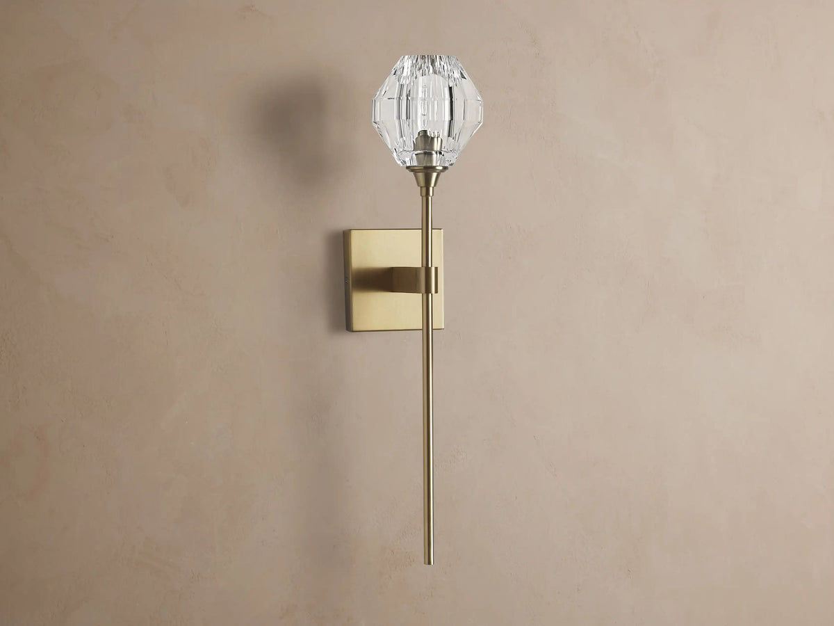 Amoroso Sconce: brass-plated wall light with long rod and illuminated faceted clear crystal shade on a brown wall.