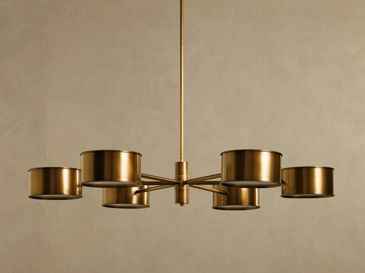 Daphne Metal Round Chandelier with 6 antique brass cylindrical metal shades radiating from a central rod, glowing softly.