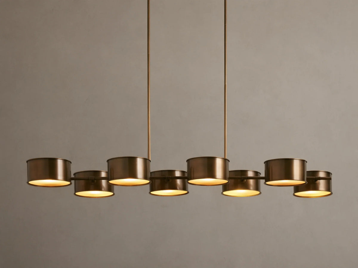 Daphne Metal Linear Chandelier with 8 warm bronze drum shades suspended from rods, emitting a soft downward glow.