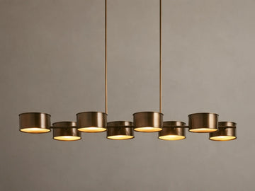 Daphne Metal Linear Chandelier with 8 warm bronze drum shades suspended from rods, emitting a soft downward glow.