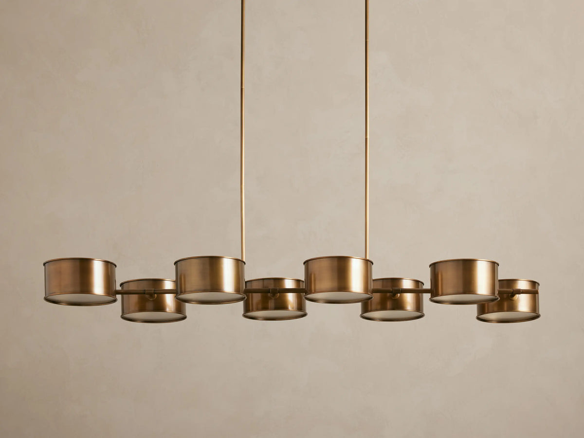 Daphne Metal Linear Chandelier with 8 warm bronze drum shades suspended from rods, emitting a soft downward glow.