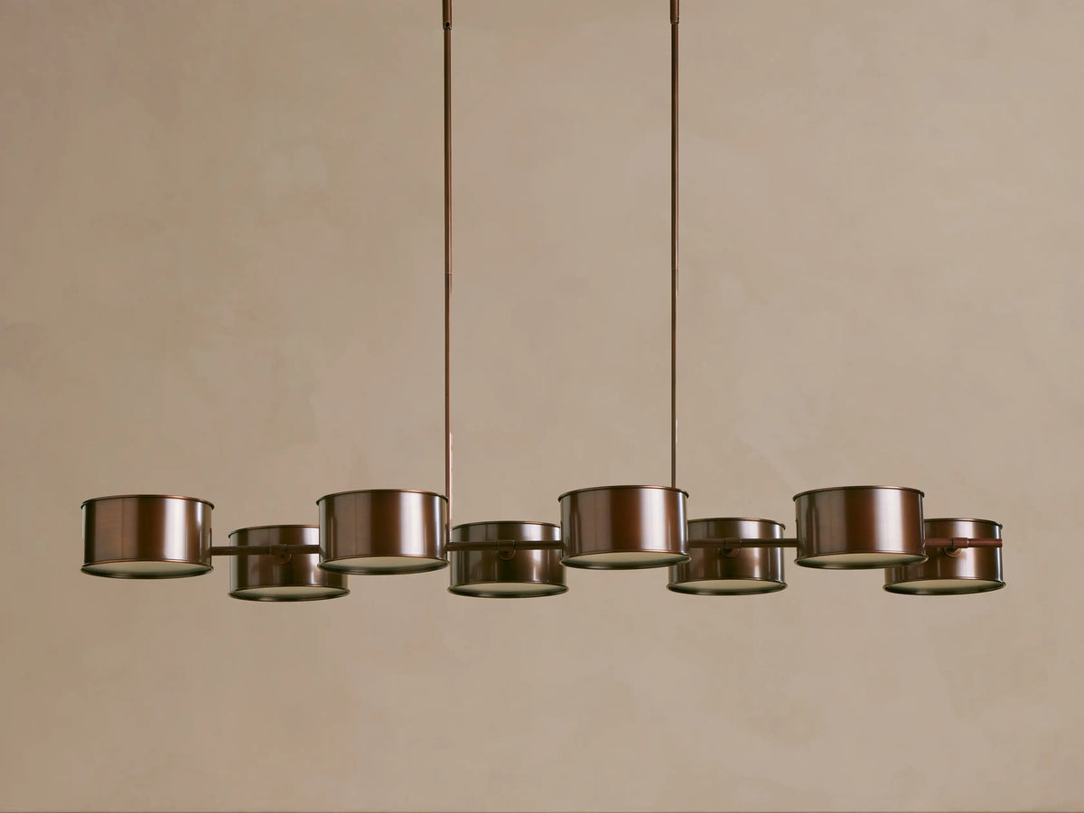 Daphne Metal Linear Chandelier, dark bronze, with 8 glowing cylindrical shades suspended from two rods against a beige wall.