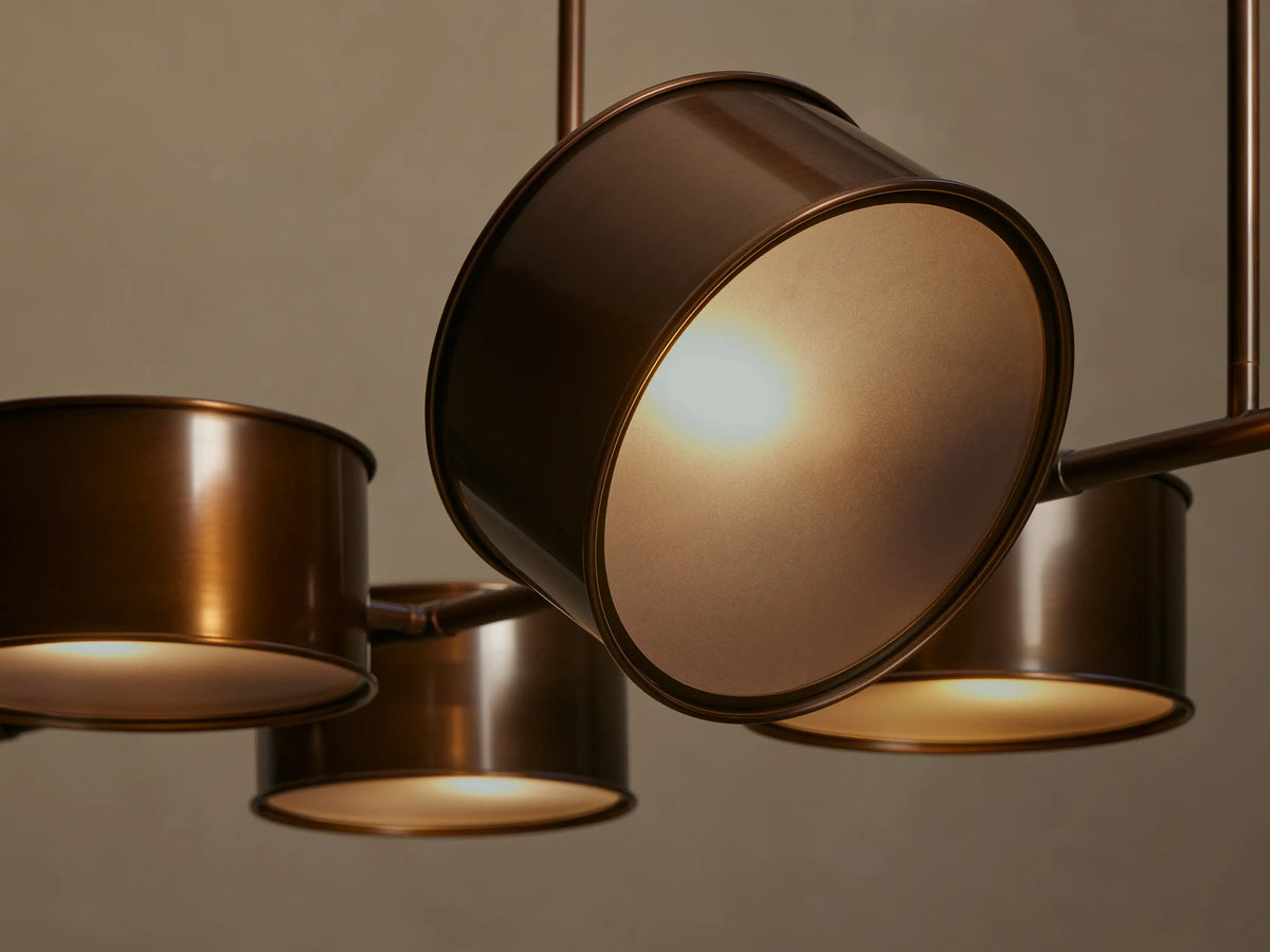 Close-up, angled view of a Daphne Metal Linear Chandelier. Warm bronze metal drum shades emit a soft, inviting yellow light.