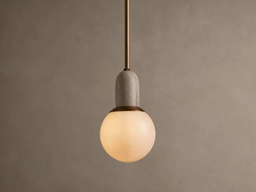 Christie Pendant light hanging. Features a brass rod, natural gray stone cap, and a glowing spherical milk glass shade.