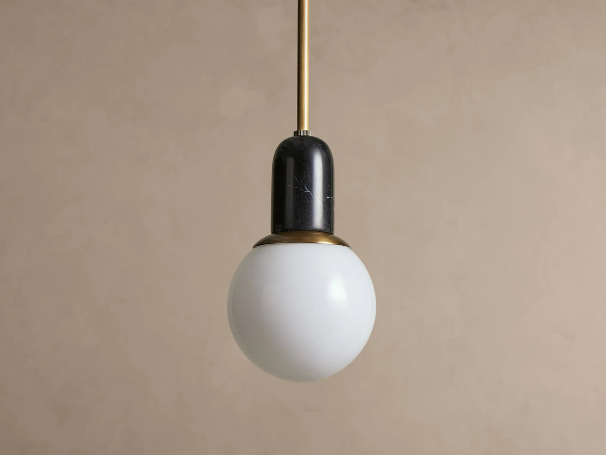 Christie Pendant light featuring a brass rod, black natural stone cap, and spherical white milk glass shade, hanging.