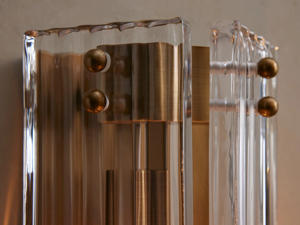 Delsie Sconce with burnished brass metalwork and fluted clear glass panels mounted vertically on a light gray wall.
