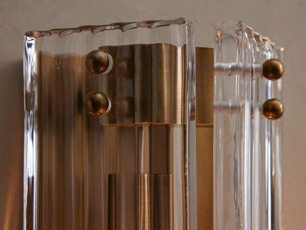 Delsie Sconce with burnished brass metalwork and fluted clear glass panels mounted vertically on a light gray wall.