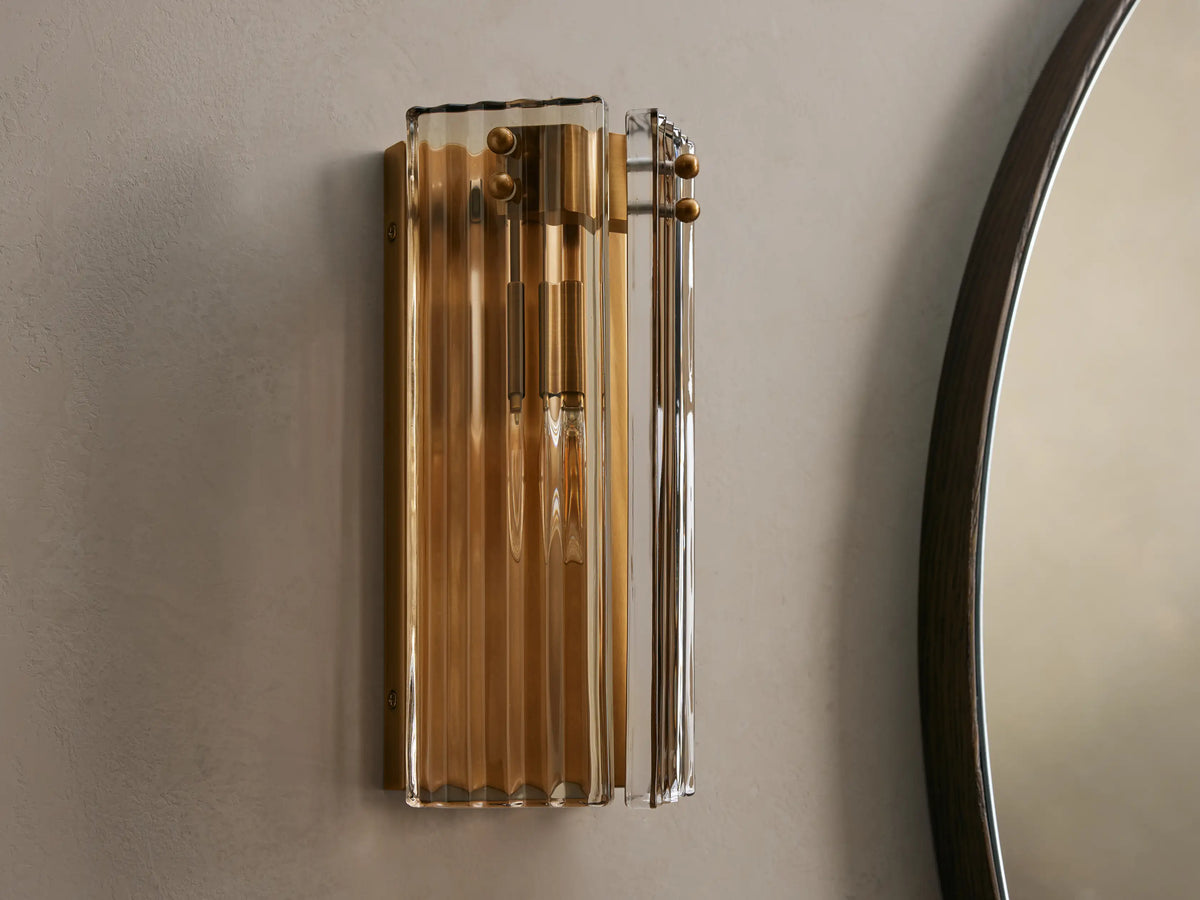 Vertical Delsie Sconce with fluted glass and burnished brass finish, glowing warmly on a grey wall next to a dark mirror.