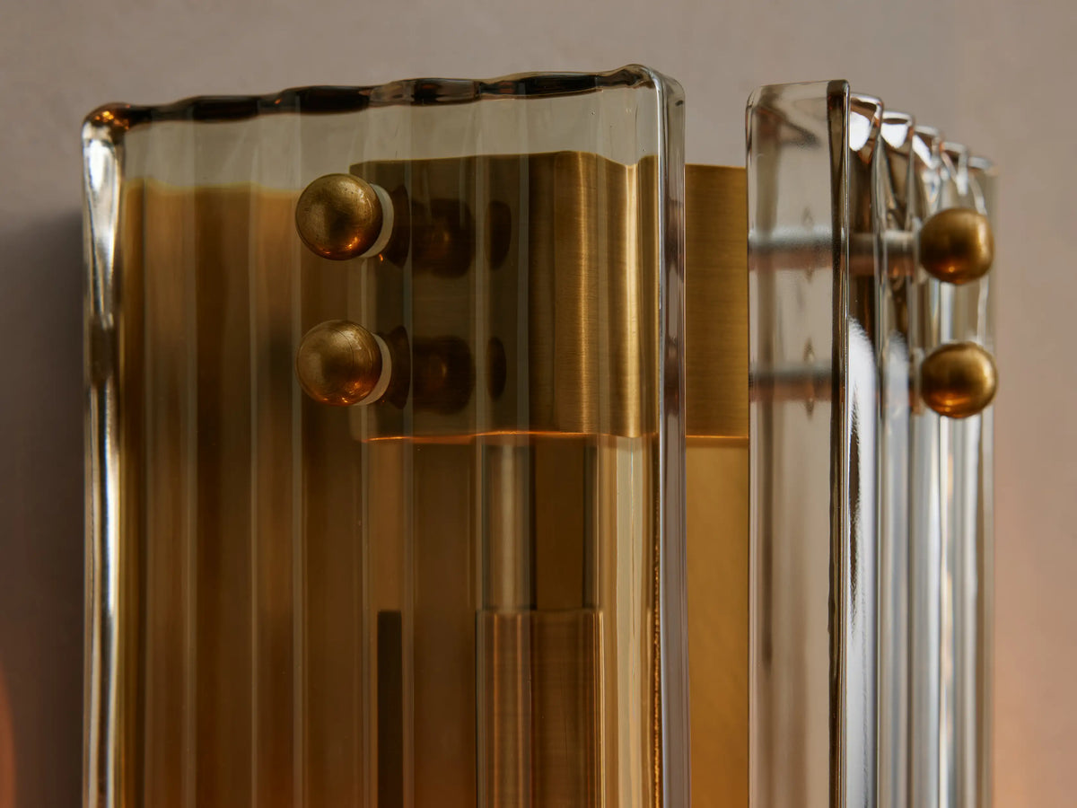 Delsie Sconce, a vertical fluted glass and burnished brass wall light, mounted on a grey wall next to a dark-framed mirror.