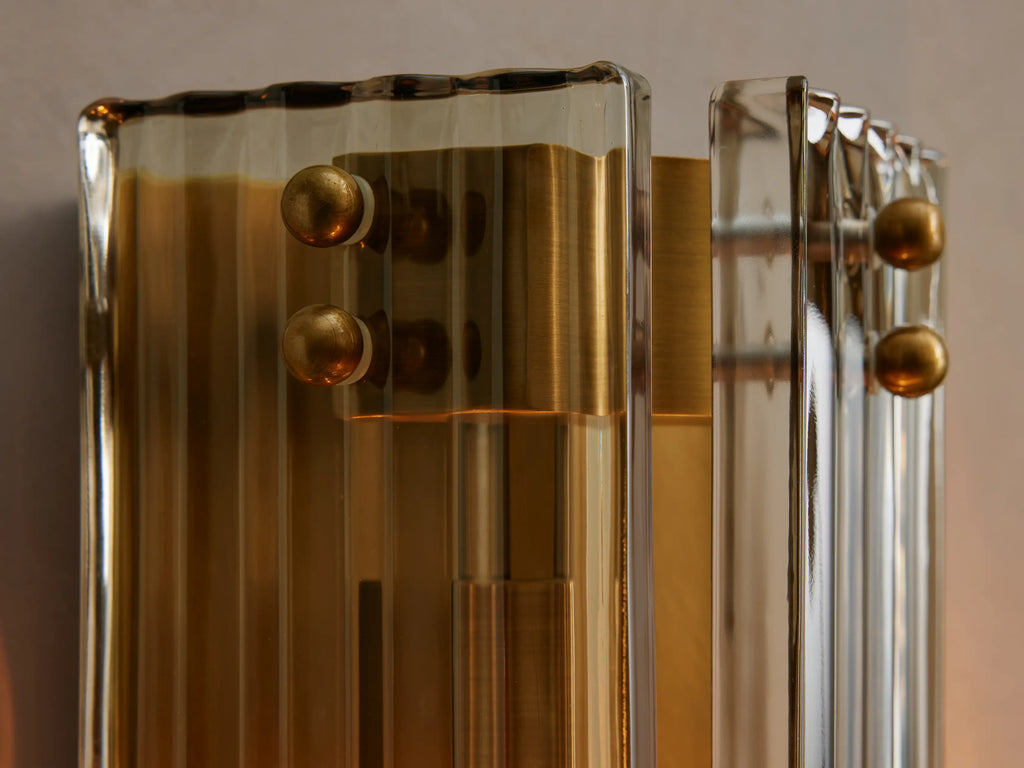 Delsie Sconce, a vertical fluted glass and burnished brass wall light, mounted on a grey wall next to a dark-framed mirror.