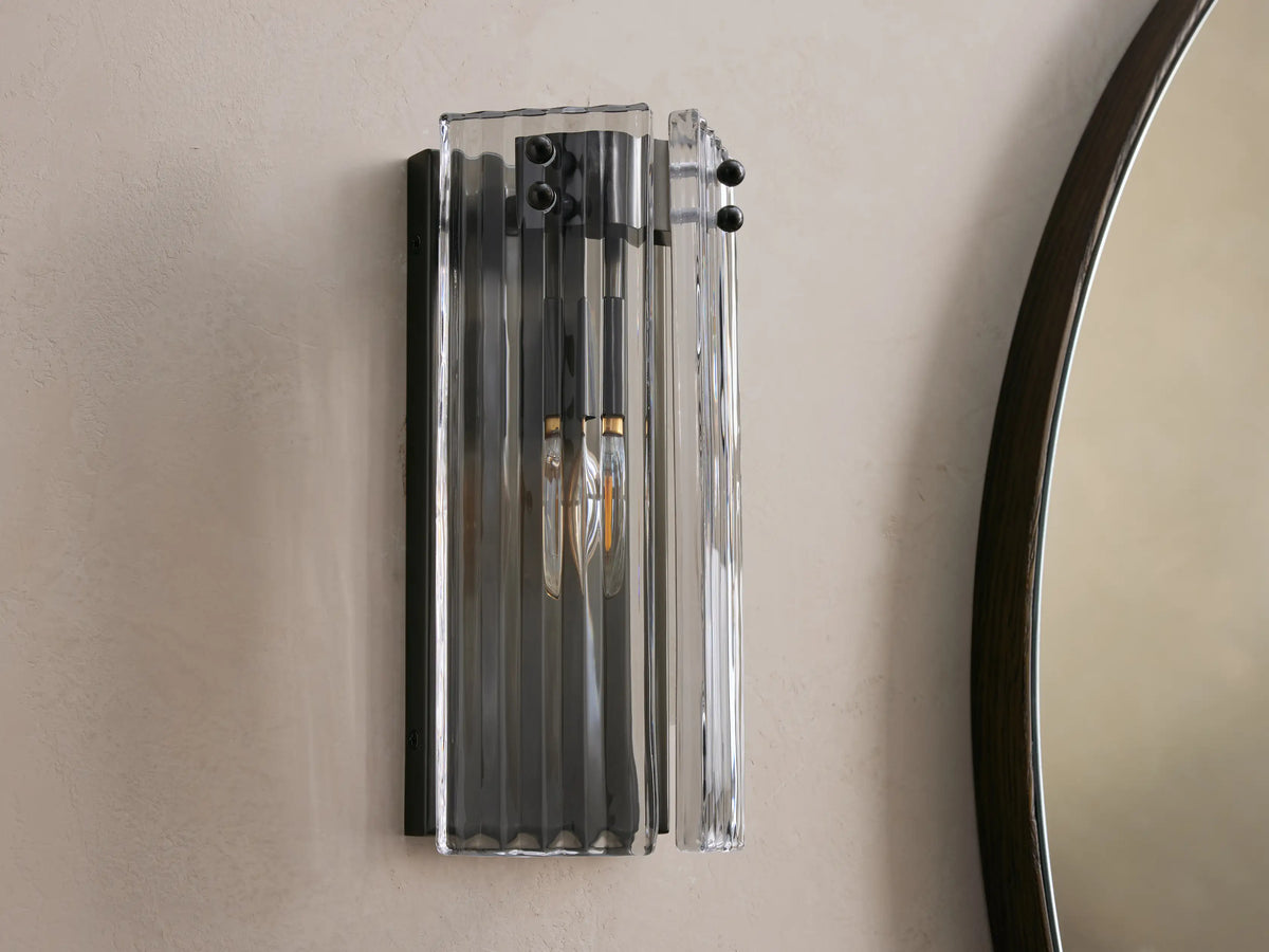 Rectangular clear fluted glass Delsie Sconce with dark metal base, glowing warmly on a beige wall next to a mirror.