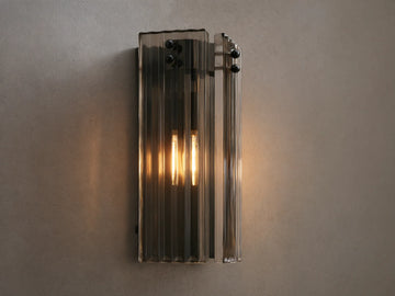 Delsie Sconce: Vertical light with ribbed smoky glass, dark metal frame, and two glowing bulbs, on a light wall beside a mirror.