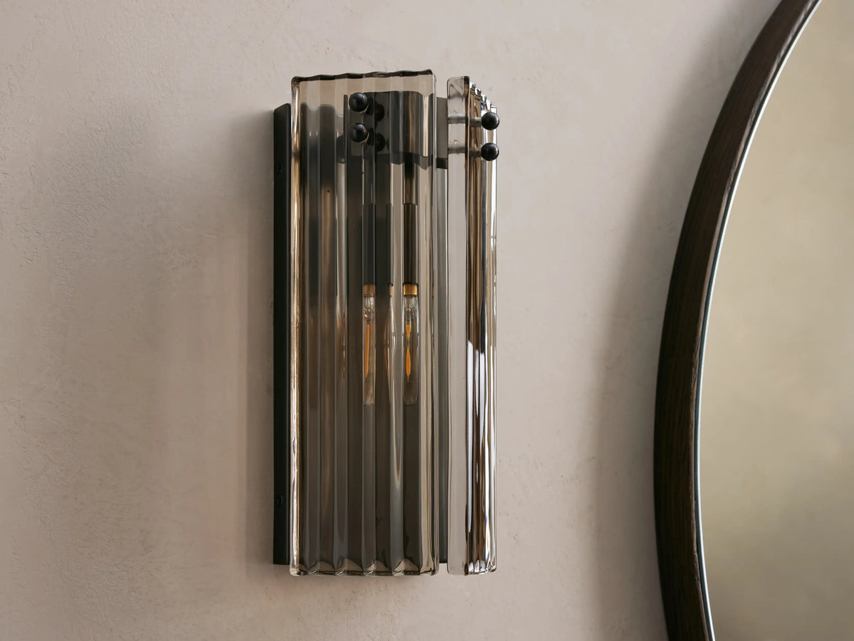 Delsie Sconce: Vertical light with ribbed smoky glass, dark metal frame, and two glowing bulbs, on a light wall beside a mirror.