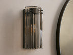 Delsie Sconce: Vertical light with ribbed smoky glass, dark metal frame, and two glowing bulbs, on a light wall beside a mirror.