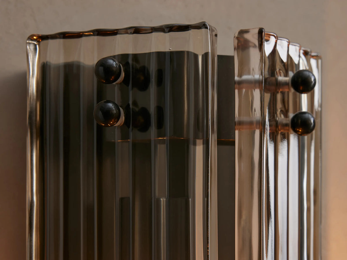Vertical Delsie Sconce with fluted gray glass panels & dark metal frame, two amber bulbs, mounted on a light wall by a mirror.