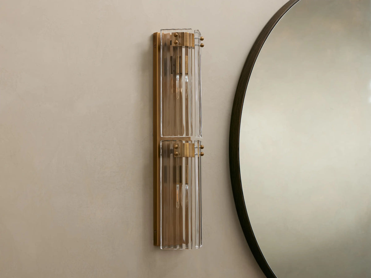Delsie Double Sconce, a vertical burnished brass and fluted glass wall light with two glowing bulbs, on a neutral wall next to a round mirror.