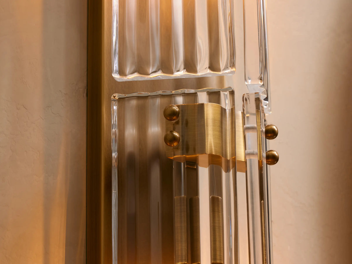 Vertical Delsie Double Sconce with burnished brass accents, two ribbed glass shades, on a beige wall beside a round mirror.