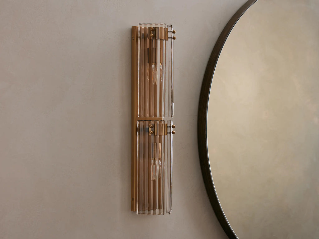 Delsie Double Sconce with burnished brass frame and ribbed glass, glowing warm, mounted vertically next to a round mirror.