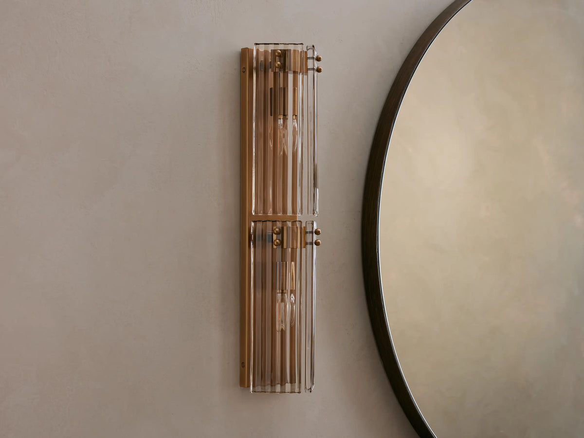 Delsie Double Sconce with burnished brass frame and ribbed glass, glowing warm, mounted vertically next to a round mirror.