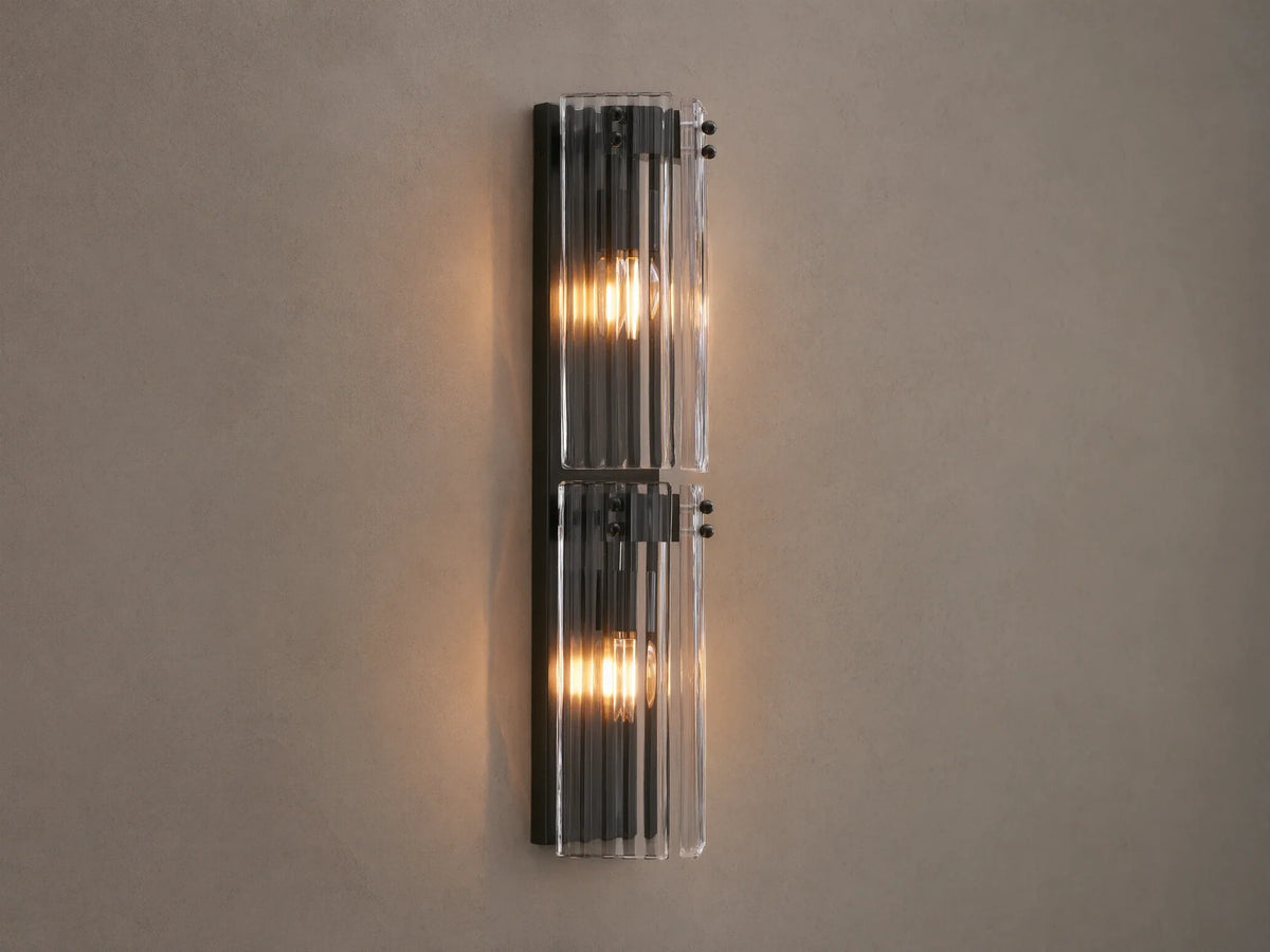 Vertical Delsie Double Sconce, dark frame with two illuminated bulbs behind ribbed clear glass, mounted on a wall near a mirror.