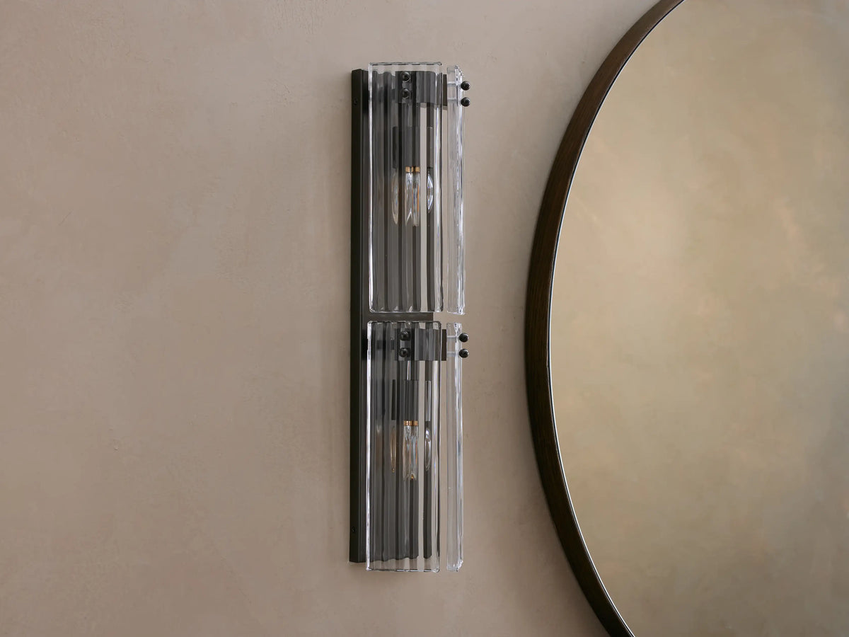 Vertical Delsie Double Sconce, dark frame with two illuminated bulbs behind ribbed clear glass, mounted on a wall near a mirror.